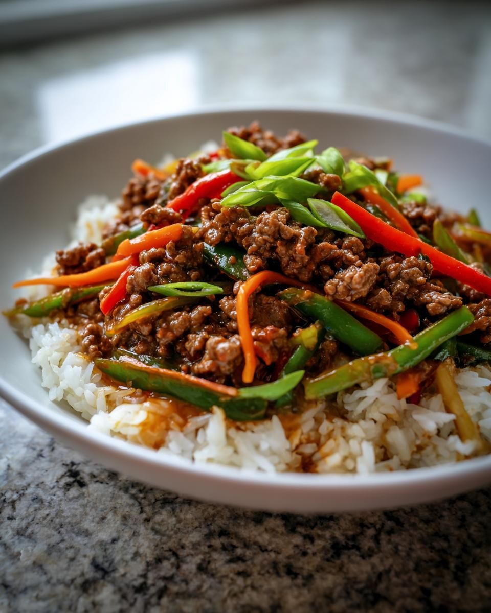 A bowl of white rice topped with a savory Asian ground beef stir fry featuring green beans, carrots, and red bell peppers, garnished with green onions.