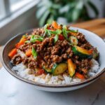 A bowl of white rice topped with savory Asian ground beef and mixed vegetables like zucchini and carrots, garnished with green onions.