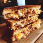 Gegrilde kaas sandwich gevuld met spek en gesmolten kaas, op een houten plank.