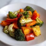 A close-up of a white plate filled with delicious roasted vegetables, including broccoli, carrots, and red peppers, seasoned with herbs.