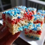 A hand holding a slice of Festive 4th Of July Rice Krispie Treats, layered with red and blue colors and topped with white frosting and star sprinkles.