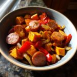 A close-up of a bowl filled with flavorful smoked sausage with sweet potato stir fry, featuring chunks of sweet potato, sliced sausage, red bell peppers, and onions.