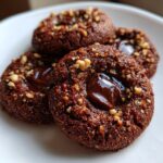 Close-up of Irresistible Chocolate Hazelnut Thumbprint Cookies filled with glossy chocolate ganache and topped with chopped hazelnuts.