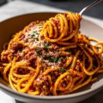 A close-up of a fork twirling Irresistible Spaghetti Bolognese, coated in a rich meat sauce and topped with Parmesan cheese and parsley.