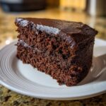 Een dikke plak Poolse chocoladetaart met glanzende chocoladeglazuur op een wit bord.