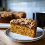 Een plak Pumpkin Coffee Cake met Streusel op een bord, met de rest van de cake en een mok koffie op de achtergrond.