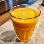 A close-up of a glass filled with a vibrant orange Papaya Orange Smoothie, topped with foam.