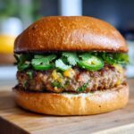 A close-up of a Spicy Jalapeno Turkey Burger on a wooden board, featuring fresh cilantro and sliced jalapenos.