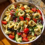 A close-up overhead view of a Vibrant Tortellini Pasta Salad Recipe with fresh ingredients like cherry tomatoes, cucumbers, olives, and herbs.