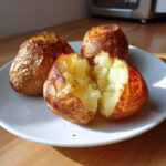 Three golden-brown Air Fryer Baked Potatoes on a white plate, one split open revealing fluffy interior.