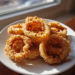 Close-up of golden, crispy Air Fryer Fried Calamari rings piled on a white plate near a window.