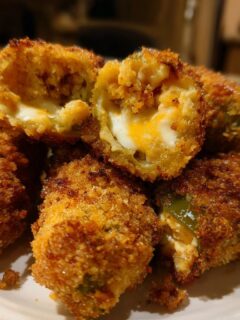 Close-up of golden brown Air Fryer Jalapeno Poppers stacked on a white plate, one is cut open showing the melted cheese filling.