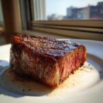 Close-up of a thick, medium-rare Air Fryer Steak with a dark, seasoned crust resting in its juices on a white plate.