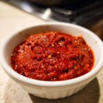 A close-up of thick, vibrant red Ajvar Roasted Red Pepper Spread in a small white bowl, showing texture and seeds.