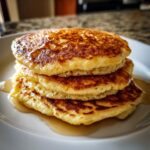 A close-up of a stack of three perfectly golden brown Almond Flour Pancakes drizzled with maple syrup on a white plate.