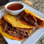Two golden, crispy tacos filled with shredded Beef Birria, served on a white plate next to a small bowl of dipping consommé.