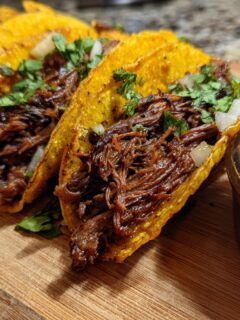 Close-up of crispy tacos filled with shredded Beef Birria, topped with onion and cilantro, served next to a bowl of consommé.