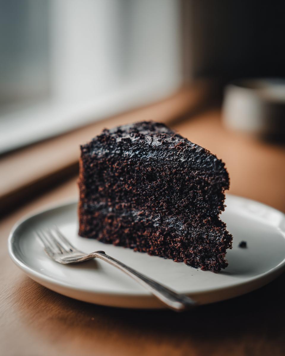A dark, moist slice of Black Magic Chocolate Cake served on a light plate with a silver fork.