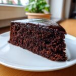 A close-up of a moist slice of Black Magic Chocolate Cake topped with shiny dark chocolate ganache.