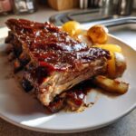 Close-up of tender Braised Apple BBQ Ribs And Potatoes smothered in thick, glossy BBQ sauce on a white plate.