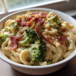A close-up of a white bowl filled with creamy Broccoli Bacon Macaroni And Cheese, topped with crispy bacon pieces.