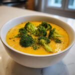 A close-up of vibrant orange Broccoli Cheese Soup topped with bright green broccoli florets in a white bowl.