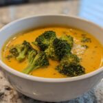 Close-up of a vibrant orange Broccoli Cheese Soup topped with fresh green broccoli florets in a white bowl.