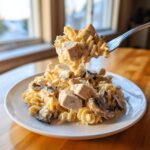A fork lifting a creamy bite of Baked Turkey Tetrazzini, showing pasta, diced turkey, and mushrooms over a plate.