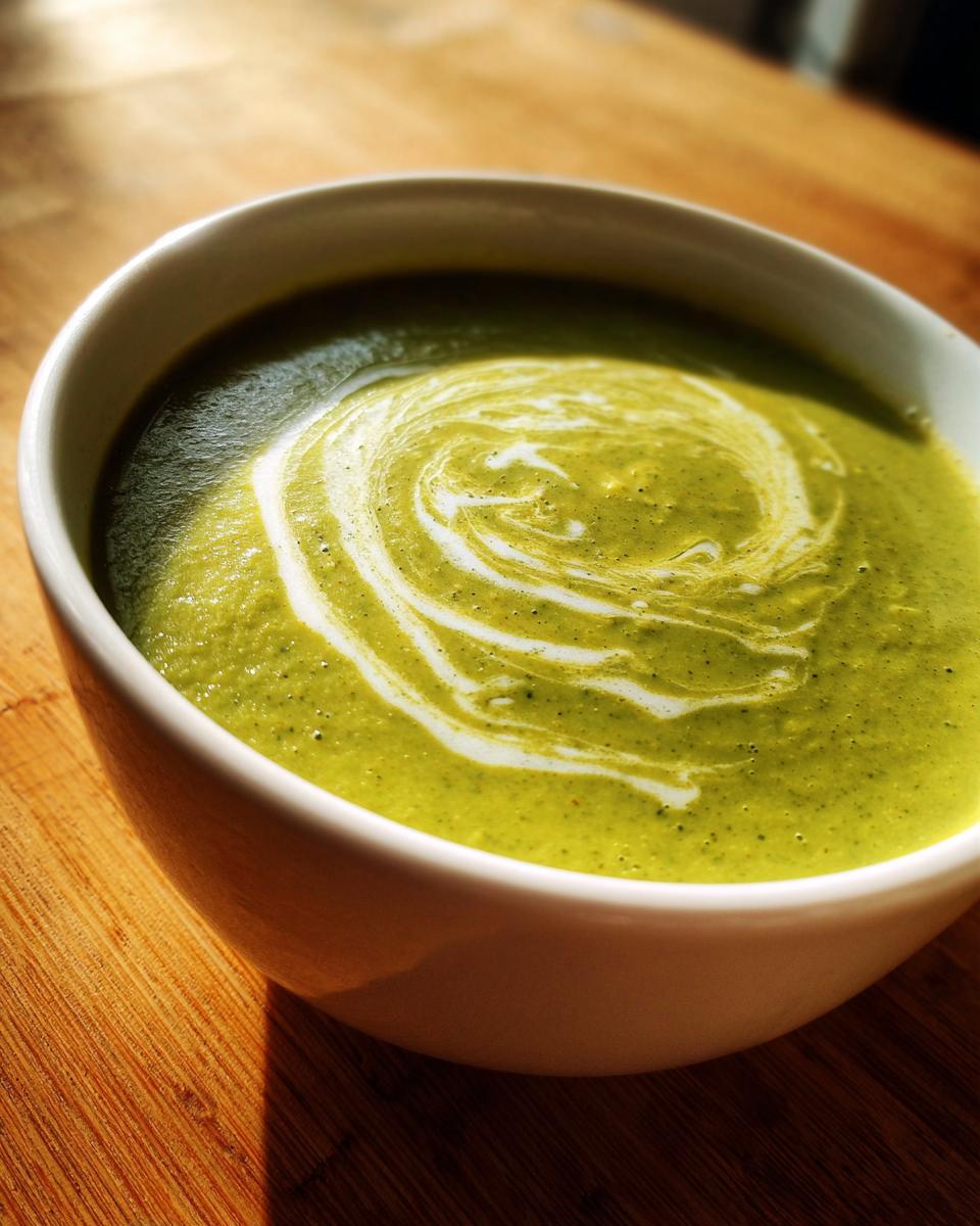 Close-up of vibrant green Broccoli Potato Soup in a white bowl, topped with a cream swirl.