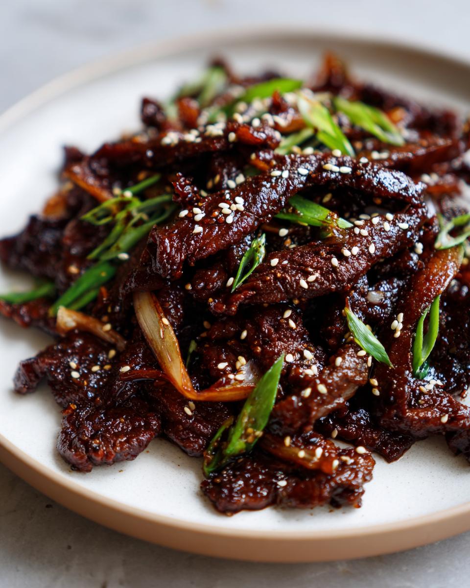 Close-up of a plate piled high with glistening, dark glazed Beef Bulgogi, garnished with white sesame seeds and sliced green onions.