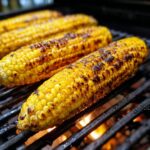 Close-up of several ears of Grilled Corn On The Cob cooking on a barbecue grill with visible char marks.