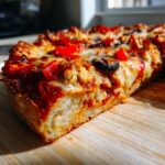 A thick slice of irresistible chicken crust pizza, showcasing melted cheese, red peppers, and olives on a wooden board.