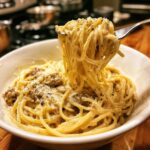 A fork lifting a swirl of Irresistible Smoked Cream Cheese Pasta from a white bowl, topped with Parmesan and black pepper.