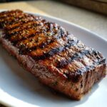 Close-up of a perfectly grilled flank steak, seasoned and juicy, on a white platter.