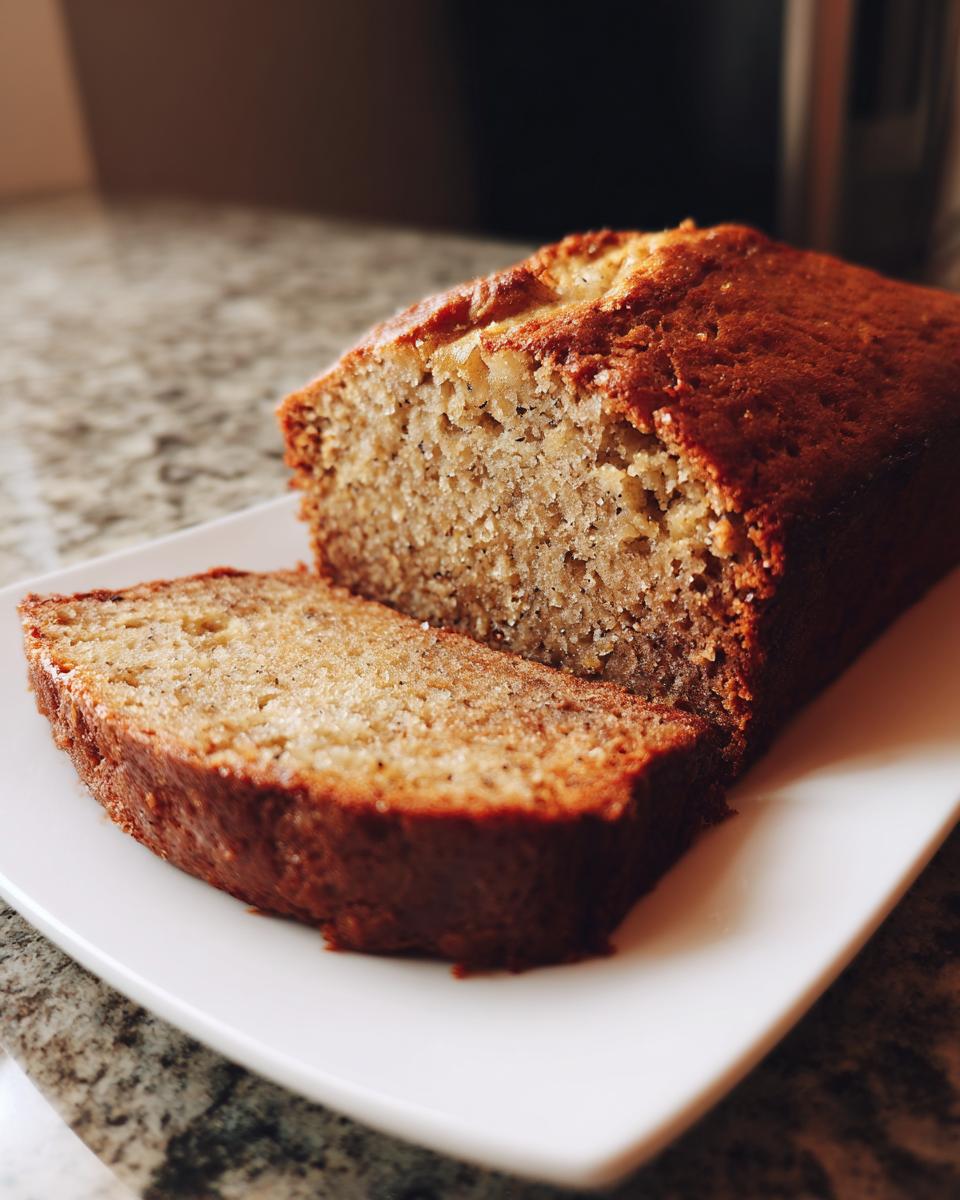 A freshly baked loaf of Banana Bread with one thick slice cut and resting against it on a white plate.
