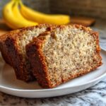 Two thick slices of moist Banana Bread resting on a white plate with whole bananas blurred in the background.