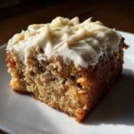 Close-up of a moist slice of Banana Cake With Cream Cheese Frosting on a white plate.