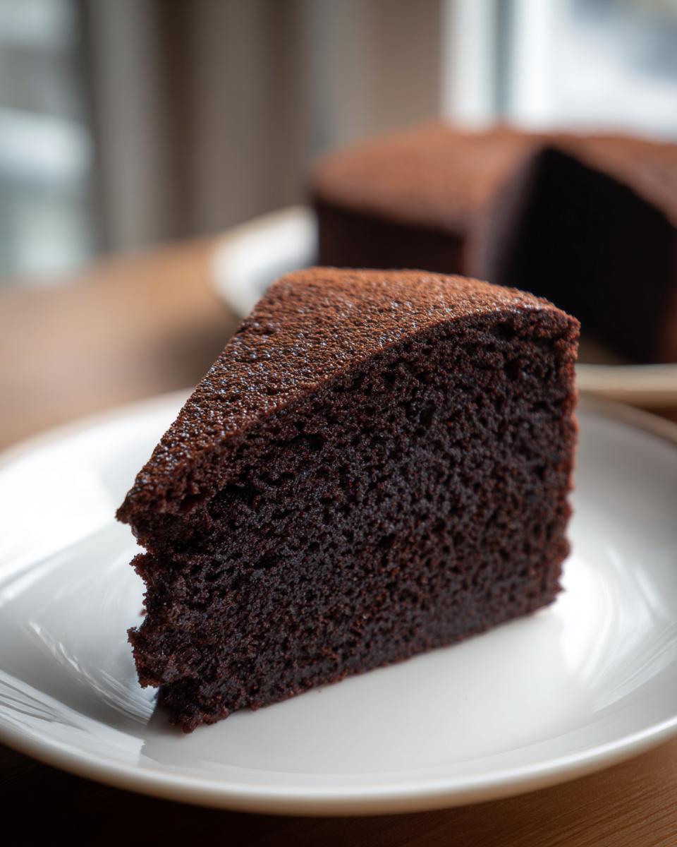 A close-up of a moist slice of Black Magic Chocolate Cake dusted with cocoa powder on a white plate.