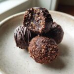 A stack of rich, dark chocolate Bourbon Balls, one bitten into showing the textured interior, resting on a light ceramic plate.
