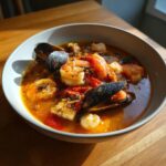 A close-up of a rich, orange-red Bouillabaisse soup filled with shrimp, mussels, and white fish chunks.
