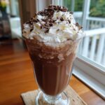 A tall glass filled with a rich chocolate milkshake, topped with a generous swirl of whipped cream and chocolate shavings. Indulge In The Ultimate Chocolate Milkshake Delight.