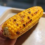 Close-up of a perfectly roasted ear of corn on the cob, with golden kernels and char marks, held in a hand.