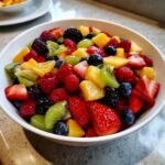 A close-up of a white bowl filled with a Vibrant Fresh Fruit Salad, featuring strawberries, blueberries, raspberries, kiwi, pineapple, and oranges.