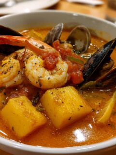 Close-up of a vibrant, orange-hued Bouillabaisse soup featuring shrimp, mussels, clams, and chunks of potato.