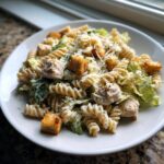 Een bord met romige Chicken Caesar Pasta Salad, met fusilli pasta, stukjes kip, croutons en geraspte kaas.