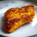 Close-up van een perfect gebakken stuk Cornflake Chicken met een knapperige, goudgele korst op een wit bord.