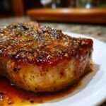 Close-up van een dikke varkenskotelet bedekt met een glanzende honing knoflook saus, geserveerd op een wit bord.