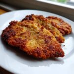 Twee goudbruine, krokante stukken Chicken Milanese met peperkorrels op een wit bord.