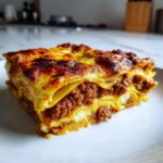Close-up van een dikke, goudbruin gebakken portie Lasagna Bolognese op een wit bord.