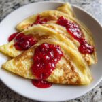 Close-up van drie gevouwen Lemon Crepes With Raspberry Sauce, rijkelijk besprenkeld met helderrode frambozensaus.